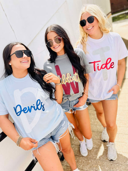 Three women wearing t-shirts with 'Devils', 'W', and 'Tide' glitter puff text, standing outdoors.