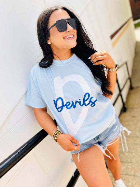 Woman wearing a light blue t-shirt with 'Devils' printed on it, standing against a white wall.